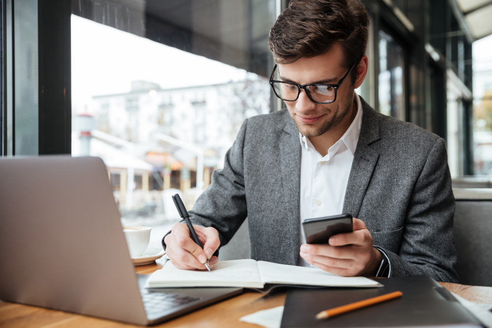 homem branco com camisa branca e blazer cinza com calculadora na mão escrevendo e um caderno